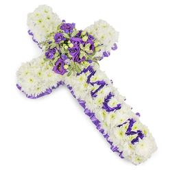 This deeply respectful floral cross, pictured against a minimalist white backdrop, is built from countless white chrysanthemum blooms, each one placed to form a soft, continuous surface that looks almost like a cloud. The edges are traced with a pleated purple satin ribbon, its ruffles introducing a refined, solemn border that lifts the eye inward. The vertical bar of the cross bears the lovingly spelled letters "MUM," formed from smaller, richly purple flower heads - likely eustoma or compact spray chrysanthemums - which stand in bright, personal contrast to the white field. Centered where the arms meet, a lush cluster of deep violet roses and pale lavender lisianthus forms the arrangement's heart: roses with velvety, layered petals and lisianthus with delicate, ruffled mouths mingle with variegated foliage whose pale margins and darker centres provide a crisp, botanical outline. Interspersed green berries and slender stems add tactile interest and depth. The photograph's soft lighting accentuates the layered purples - from almost-blue violet to whispering lavender - and the chrysanthemums' cushion-like texture, conveying both dignity and intimacy. As Clerkenwell Flowers, preparing tributes for services around Exmouth Market, St John Street and the Barbican, we find this design expresses quiet affection and respectful mourning; it reads well at a memorial, a funeral chapel, or laid beside a coffin. The scent is faintly floral and clean, the composition steady and consoling - a piece intended to carry a family's tenderness and farewell.