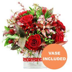 A full, romantic bouquet in rich shades of red and green is showcased against a pure white background, much like a statement piece ready to be sent from Clerkenwell to a hidden courtyard apartment near St John Street. The central focus is a cluster of sumptuous red roses, their velvety petals spiralling inward, some buds still tight with promise, others opening just enough to reveal their intricate, layered hearts. Nestled between these classic blooms are smaller deep red flowers, akin to alstroemeria or similar multi-flowered stems, which introduce a sense of rhythm and movement to the design. Delicate sprays of tiny blush-pink and white filler blossoms, suggestive of waxflower or limonium, weave gently through the arrangement, adding a light, airy contrast against the intense reds. Surrounding this core of flowers is an abundant frame of foliage: broad, glossy leaves mingle with finer, fresher green sprigs to create a lush, rounded shape that feels carefully composed yet natural. Accents of burgundy and reddish-brown leaves, some with discreet variegation, provide subtle shadows and depth, like the warm tones you might glimpse in the evening light along Clerkenwell Green. The bouquet is presented in a sleek, clear glass vase with a modern rectangular base, allowing the stems to be seen and giving the whole piece a polished, contemporary feel. A red ribbon decorated with evenly spaced white heart outlines wraps around the vase, reinforcing the romantic theme with a playful nod. In the bottom right corner, a bold circular orange label reads "VASE INCLUDED" in white uppercase text, highlighting that this is a complete, gift-ready floral arrangement ideal for love notes, anniversaries, or special deliveries across EC1.