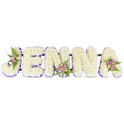 There is a quiet artistry to this funeral tribute: the name JENNA formed in large, three-dimensional letters composed of hundreds of small, ivory-white chrysanthemums or closely related florets, arranged against a pristine white field so that each letter feels soft, voluminous, and serenely luminous. A subtle purple trim-appearing as a narrow ribbon or a halo of tiny lavender blooms-outlines the inner contours of each letter, offering contrast and gentle definition that guides the eye across the name. Thoughtful floral accents punctuate the design: a cluster of blush-pink and pale lavender spray roses nestles into the curve of the J, while another compact grouping of cream-toned roses and sprays of lavender rests along the second N, and a final arrangement graces the upper right of the A. Dark-green fern fronds cradle these clusters, their fine texture balancing the chrysanthemums' dense, cloud-like surface. The roses' soft scent mingles faintly with the greener, slightly earthy tones of the foliage; overall the display reads as tactile and comforting rather than ostentatious. Narrow, unobtrusive dark bars connect the letters along their bases, giving the tribute structure and making it suitable for placement by the chapel at a funeral or at a wake. Designed with the sensitivity of a London florist who knows Clerkenwell's quieter corners-near Saint John's Street and Clerkenwell Green-this arrangement offers a personal, dignified way to honour someone's name with calm beauty and thoughtful detail.