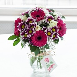 A lush, professionally styled bouquet fills a compact, clear glass vase, set against a softly diffused white backdrop that feels like a bright Clerkenwell loft or studio on an overcast morning. The arrangement is dominated by striking gerbera daisies in deep fuchsia and magenta tones, each bloom centred with a dark, almost black disk that gives depth and drama. Around them, bicolour chrysanthemums unfurl petals brushed in vivid purple at the tips, fading gracefully into white before meeting warm yellow centres, creating a rhythmic pattern of colour. Creamy white roses, both in full bloom and in elegant bud form, are tucked between the bolder flowers, lending a classic, timeless charm that balances the modern intensity of the gerberas. Wisps of white baby's breath weave gently through the bouquet, their tiny blossoms like scattered stars adding lightness and movement, while rich green foliage anchors the whole design and frames the colours. The stems are neatly gathered in a squat, jar-style vase whose clear, textured glass reveals the fresh, cleanly cut stems and the water they drink. A length of rustic twine is tied around the vase's neck, forming a simple bow from which a small rectangular tag hangs, printed with a delicate pink floral emblem and flowing script, suggesting a thoughtful handwritten note. This bouquet could easily be imagined waiting on a table in a Clerkenwell townhouse near St John Street, ready to greet someone returning home, its vibrant magentas, purples, and creams radiating warmth, celebration, and the comforting sense of flowers chosen with care by a knowledgeable local florist.