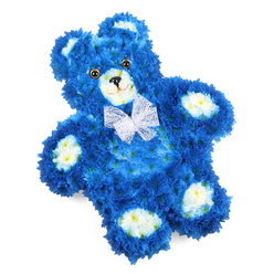 Set against a crisp white backdrop, this floral teddy bear tribute looks as though it has been sketched from childhood memory and then brought to life with flowers. The bear's rounded body, head, and limbs are densely carpeted in royal blue blossoms, each small petal tucked neatly into the next to imitate plush fur. The saturation of the blue is deep yet calming, evoking a sense of serenity that is especially moving for sympathy work. Creamy white and pale buttery yellow blooms are carefully placed to shape the bear's muzzle, inner ears, and oval paw pads, adding definition and warmth to its features. Dark, shiny button-style eyes and a black triangular nose are positioned centrally, with a fine curved line beneath to suggest a soft, reassuring smile. Around the neck sits a sheer white mesh or organza bow, its delicate, ruffled loops catching the light and echoing the gentle theme of innocence and care. The composition is photographed straight on, with the bear lying flat yet appearing almost three-dimensional due to the layering of petals and the subtle shadows between blooms. From my perspective as a local Clerkenwell florist, this design feels especially suited to heartfelt farewells and baby memorials at churches and venues around Farringdon Road, Clerkenwell Green, and nearby gardens, where families often seek a tribute that is both tender and distinctive. The royal blue hue can symbolise peace, protection, or remembrance for a baby boy, while the teddy bear shape recalls comfort and childhood security. It is a piece that quietly offers solace, whether placed beside a casket, on a memory table at a wake, or as a loving keepsake arrangement delivered to a home in EC1.