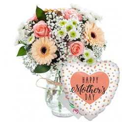 Set against a crisp white background, this Mother's Day scene captures the feeling of walking into a calm Clerkenwell studio filled with the scent of fresh blooms. At the heart of the image, a clear glass vase holds a generous, rounded bouquet, its transparency revealing clean stems neatly gathered by an experienced florist's hand. Soft peach-pink roses, fully open yet still tidy in their spiral, form the romantic core of the design. Beside them, bold peach gerbera daisies with deep chocolate-brown centres add a playful, almost sunny character, like a patch of light breaking through the clouds over nearby Exmouth Market. Between these focal blooms, clusters of delicate white gypsophila create a lace-like veil, while petite white chrysanthemums with light green centres add volume and a touch of botanical precision. The foliage, in various shades of fresh green, peeks strategically from behind petals and around the rim of the vase, giving the bouquet a natural, just-picked energy. Around the vase neck, a slim twist of rustic twine suggests a handcrafted, neighbourhood touch that feels typical of Clerkenwell's creative side streets. Tucked modestly behind the flowers is a small white card, its presence hinting at personal words of love rather than drawing attention away from the blossoms. In front and slightly to the right, a heart-shaped Mylar balloon rests against the arrangement, its white surface dotted with shimmering gold and silver circles. Centred on the balloon, a soft peach heart frames the message "HAPPY Mother's DAY" in bold black script, the sweeping letterforms mirroring the gentle curves of each petal. The entire composition evokes tender gratitude and quiet celebration, ideal for local Mother's Day deliveries around Clerkenwell and Farringdon.