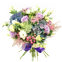Lush, elegant, and evocative of Clerkenwell's artistic heart, this floral bouquet is thoughtfully arranged by local florists to mirror the lively tapestry of this London neighbourhood. The arrangement bursts with multi-petaled hydrangeas in saturated indigo, rich teal, and subtle purple-colours that nod to the creative energy along Clerkenwell Road. Clustered close, delicate lavender and cream roses bloom with classic poise, their soft petals unfurling as gracefully as the morning light over Charterhouse Square. White ranunculus, tightly layered and full, bring a gentle textural contrast, while pale yellow lisianthus blooms are scattered throughout, echoing the delicate beauty of springtime along St James's Walk. Among the larger flowers, vibrant fuchsia fillers flash, their small blossoms creating joyful pops of colour, while airy pale pink astilbe feathers the edges for a sense of playfulness. Pale green and white lacy fillers lend an ethereal, almost mist-like effect, capturing the essence of Clerkenwell's blend of new and old. Deep green foliage frames the bouquet, enhancing its freshness and design, each stem gathered and tied with a wide, sumptuous purple satin ribbon for a final flourish. The bouquet exudes joy, romance, and polished refinement-apt for celebrations, heartfelt gifting, or brightening a Clerkenwell home or office. It's a testament to local creativity and expert craft, available for seamless delivery throughout the area, from Farringdon to the City fringe.