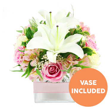 A compact yet opulent floral display is captured against a bright white backdrop, the kind of arrangement you might find ready for delivery from a trusted Clerkenwell florist just off Exmouth Market. Two pristine white Oriental lilies form the focal point, their trumpet-like blooms fully open and slightly tilted, each petal edged with a soft, natural curve that frames the prominent dark reddish-brown stamens. Around them, layers of pink roses in varying stages of bloom create a sense of movement and tenderness, from tightly furled buds suggesting new beginnings to fully opened blossoms with intricate, swirling centres that speak of romance and gratitude. Interwoven among the roses are graceful sprays of pink and white alstroemeria, some petals streaked with golden yellow and fine burgundy flecks, adding a painterly detail that rewards a closer look. Wisps of white baby's breath drift between the primary flowers like tiny clouds, softening the transitions and giving the bouquet a light, ethereal quality. A rich assortment of green foliage - broad, glossy leaves, ferny fronds, and slimmer blades - emerges from beneath the blooms, grounding the design and evoking the freshness of a morning walk through nearby Clerkenwell Green. All of this is gathered neatly into a clear, square glass vase, its transparency revealing the carefully trimmed stems and clean water inside. A pale pink satin ribbon encircles the middle of the vase in a flat band, suggesting a finished, gift-ready piece rather than a casual bunch. In the lower right corner, an orange circular graphic with bold white capitals clearly announces VASE INCLUDED, drawing the eye without distracting from the flowers. The arrangement radiates a sense of gentle joy, well-suited to mark intimate celebrations, send get-well wishes to a local clinic, or brighten a design studio near Farringdon.