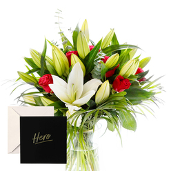Clerkenwell Flowers presents a portrait of sophistication and emotion in this bouquet, carefully composed to evoke admiration and comfort. The white lilies rise at the center, large trumpet shapes with creamy, pointed petals that feel like beacons in a refined setting. Surrounding them, green spears and curling foliage create a sculptural frame, their gloss catching the light and adding a tactile, almost velvet texture to the design. Red roses cluster throughout, their deep, ruby hue and sweet fragrance mingling with the lilies in a dewy, hypnotic aroma. Unopened buds pepper the arrangement, promising beauty still to come and hinting at ongoing celebration. The bouquet rests in a clear vase, its glass catching the daylight so each stem and leaf is outlined with precise clarity. The overall composition reads as a modern classic-ordered, generous, and deeply sincere. A card marked Hero accompanies the bouquet, making it an ideal expression of thanks, remembrance, or quiet support for someone whose spirit inspires others. Clerkenwell Flowers offers same-day delivery across Clerkenwell, Islington, and the City, bringing this refined gesture to homes, hospital rooms, and busy workplaces alike. The scent, the light, and the careful balance of bloom and greenery all say: you are seen, you are valued, you are remarkable.