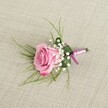 A single blush pink rose boutonniere lies carefully posed on a softly textured, light beige linen cloth, as if resting on a freshly pressed jacket laid out in a Clerkenwell townhouse before a celebration. The photograph is taken from a gently elevated angle, allowing every curve of the semi-open rose to be seen: velvety petals spiral from a pale, powdery pink at the heart to a slightly deeper, more romantic pink at the outer, gently ruffled edges. Around the base of the rose, airy clouds of creamy white baby's breath cluster in delicate sprays, their tiny round blossoms catching the light and creating a soft halo that makes the central bloom feel even more luxurious. Fine, grass-like strands of fresh emerald green foliage fan lightly from behind the rose, adding a sense of movement and airy texture, while a broader, deeper green leaf has been neatly folded to cradle the stems, giving the design a tailored, almost architectural finish. The stems are bound with a satin ribbon in harmonious tones of lavender-pink and subtle magenta, the sheen of the fabric echoing the silk lapels of a groom's suit ready for a wedding near Exmouth Market or St John Street. The ribbon is tied in a precise, modest knot, suggesting the steady hand of an experienced florist. Soft, even lighting falls across the scene, revealing the linen's gentle weave and the boutonniere's intricate craftsmanship without harsh shadows. The overall mood is tender and romantic, a timeless finishing touch for a groom, groomsman, or prom guest walking the streets of Clerkenwell on a day filled with quiet anticipation and celebration.