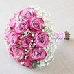 A luxurious round bouquet, skillfully crafted by florists in Clerkenwell, features a captivating display of two-toned pink roses nestled among airy clouds of white baby's breath. The roses are the star: their tightly furled centres blush in soft lavender-pink, gradually deepening into radiant fuchsia and magenta along the edges, creating mesmerizing visual depth. Each rose is at the peak of freshness, arranged in a harmonious, domed cluster of a dozen blooms. Dainty tufts of baby's breath are woven throughout, offering a light, romantic accent that seems to float between the vibrant petals. The stems are neatly gathered and wrapped in a silky pink ribbon, its sheen echoing the softer tones of the flowers, while a spiralled strand of white pearls brings a refined, celebratory touch-ideal for bridal bouquets at local venues like Charterhouse Square. The bouquet sits atop a pale, textured fabric that sets off the bold colours, illuminated by gentle daylight that flatters every petal. Hints of delicate rose and fresh greenery waft from the bouquet, evoking feelings of love, elegance, and sentiment-perfect for weddings, engagements, anniversaries, or as a statement arrangement for intimate Clerkenwell gatherings.