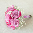 A romantic bridal-style bouquet of bi-colour roses and baby's breath lies on a pale, woven beige fabric, its diagonal placement drawing the eye from the lush blooms at the top left down to the pearl-wrapped stems at the bottom right. The roses are striking yet gentle, each bloom showing a gradient from cool lavender and light mauve in the centre to a vivid fuchsia and magenta pink along the petal edges, creating a watercolour-like effect. Their petals curl and layer in soft spirals, giving the arrangement depth and texture. Threaded generously around these feature roses, delicate sprays of white gypsophila - commonly known as baby's breath - form airy clouds, their tiny round blossoms adding a sense of innocence and softness to the composition. The stems are bound with a rich purple satin ribbon, smooth and slightly glossy, then adorned with a spiralling line of small, white faux pearls, like a bracelet wrapped around the handle. This detail gives the bouquet a polished, occasion-ready feel, perfect for a wedding at the Town Hall or a civil ceremony near Clerkenwell Green. The neutral background fabric has a subtle, tactile weave that keeps the focus on the florals while suggesting a natural, relaxed setting, perhaps a linen tablecloth in a local Clerkenwell venue. The overall impression is one of careful craftsmanship and understated luxury, an arrangement that feels equally suited to a bridesmaid's bouquet, a prom posey collected on the way from Farringdon Station, or a special anniversary gift delivered to a home overlooking Exmouth Market.
