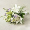 A lush and sophisticated bridal bouquet rests on a subtly textured, light beige linen-an elegant touch that could belong to any Clerkenwell loft or historic townhouse. The composition is dominated by magnificent, trumpet-like white Oriental lilies, some fully open to display pristine petals and golden anthers, others gently unfurling in anticipation. Interlaced within this serene white canvas are clusters of lisianthus, their ruffled petals ringed with vibrant violet, and the spires of deep purple Veronica, providing depth and contemporary flair. Wispy sprigs of gypsophila scatter amidst the arrangement, their airy presence balancing the drama of the lilies and invoking the soft, clouded skies above Farringdon. Framing the entire arrangement, abundant green foliage, including delicate asparagus fern and glossy broad leaves, evokes the abundance of local flower markets and Clerkenwell's garden squares. The bouquet's stems are hand-tied and bound with an understated white ribbon, signifying attention to detail and ready for ceremonies across Clerkenwell and nearby Barbican. Touched by soft, even daylight, every petal, leaf, and floret gleams with fresh vitality, making this bouquet a timeless symbol of purity, joy, and love, expertly crafted for unforgettable wedding moments in London EC1.