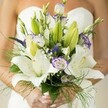This enchanting bridal bouquet radiates the artistic spirit of Clerkenwell, lovingly crafted for a bride whose hands-manicured and adorned with light polish-embrace both tradition and romance beneath a classic white wedding gown. Softly diffused natural light reveals the floral artistry: large white Oriental lilies, open and fragrant, their spotted petals illuminated and stamens glowing gold, are paired with lisianthus-each bloom edged in gentle lavender, with scattered unopened buds for a touch of mystery. Wispy sprays of green asparagus fern curve gracefully, mingling with airy baby's breath and vivid spikes of royal purple veronica, echoing the creative contrasts found along Clerkenwell Road. Lush green leaves and unopened buds provide depth against the blurred canvas of silk fabric, focusing the eye on the freshness of each flower. The entire composition exudes airy sophistication, balancing modern urban style with a sense of enduring love, perfectly suited for ceremonies and celebrations across the local Clerkenwell neighbourhood, where history and contemporary life blossom together.