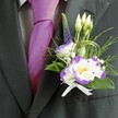 A close-up photograph shows the upper torso of a man dressed for a refined wedding or special event in Clerkenwell, the focus drawn sharply to his elegant buttonhole. He wears a perfectly tailored charcoal grey suit jacket with a defined lapel, layered over a crisp light grey checkered shirt that adds a quiet, geometric pattern. At his throat sits a glossy magenta silk tie, precisely knotted so the rich colour pools like satin in the centre of the frame, echoing the lively energy of a London celebration near Exmouth Market or St John Street. Pinned to the left lapel, the boutonniere becomes the visual heart of the image: a cluster of white lisianthus blooms edged in deep violet, their ruffled petals opening around warm yellow centres, almost glowing against the dark fabric. Around them, smaller lisianthus buds remain gently closed, promising fresh life and continuity, while a slender spire of indigo-purple veronica stretches upwards, lending vertical elegance. Wisps of fine greenery and feather-light sprigs create a soft, meadow-like texture, interspersed with tiny white filler flowers reminiscent of baby's breath. Loops of decorative green grass or wire add a contemporary twist, curving gracefully around the flowers like a subtle flourish of calligraphy. At the base, a neat white satin ribbon is tied into a tidy bow, its sheen catching the light and reinforcing the sense of tradition and care. The overall impression is one of poised, modern romance, as though the wearer is about to step from a Clerkenwell townhouse onto cobbled streets, carrying the fragrance and symbolism of a thoughtfully crafted wedding boutonniere close to his heart.