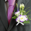 This boutonnière sits just above the heart, a compact composition that speaks of considered taste and lively sentiment. At its centre, a white lisianthus edged with a clear violet-purple reads like a delicate brushstroke, petals soft and slightly ruffled, inviting a tender touch; nearby, a crisp white dianthus with purple-ruffled margins gives a spirited, textured compliment. The arrangement is framed by rich green foliage with an earthy sheen and airy loops of ornamental grass that add motion and a sense of garden-fresh spontaneity. Small clusters of gypsophila scatter like soft confetti, brightening negative space with tiny white pinpoints, while an upright stem of deep indigo veronica introduces a vertical line that lifts the design. A neat white ribbon secures the stems, its clean finish offering a quiet note of formality against the dark suit and the luminous magenta tie beside it. Gentle light skims the petals and highlights the buds that still hold their promise, producing subtle shadows that enhance the bouquet's depth and tactility. As a Clerkenwell florist would appreciate, this boutonnière feels at home at occasions around Farringdon and St John Street-suitable for a groom stepping out from a nearby chapel or for an honoured guest arriving from Leather Lane Market. The scent is soft and garden-like, the textures balanced to remain comfortable throughout a long celebration. It's an elegantly personal detail, made with steady hands and neighbourhood knowledge that values both beauty and durability.