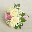 Resting elegantly on a finely woven, pale beige linen, this small floral bouquet captures the romantic heart of Clerkenwell celebrations. At the focal point, a handful of creamy roses-petals lush, full, and tenderly unfurled-are complemented by a bold magenta rose tucked just off the centre, its colour reminiscent of Clerkenwell's hidden pockets of art and creativity. Delicate baby's breath cascades among the blooms, lending a light, frothy texture and a sense of classic refinement. Dark green leaves frame the bouquet's silhouette, creating a vivid contrast to the softer flower tones and echoing the fresh ambience of local squares and gardens. The handle, visible at the edge, is tightly bound in a pink satin ribbon, further wrapped with a spiral of petite white pearls that catch the gentle daylight-an elegant detail that could easily grace a wedding at a Clerkenwell register office or an intimate soirée nearby. The bouquet's design feels both airy and intensely romantic, with every element thoughtfully crafted to evoke joy, elegance, and the artistry of a local florist devoted to making each celebration in Clerkenwell truly special.