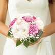 This photograph shows an elegant bridal bouquet carefully cradled, its design centred around lush, creamy peonies that glow softly in the light, like small ivory clouds. Each bloom is full and cushiony, with ruffled petals layered so densely that they almost seem to ripple, giving the whole arrangement a sense of luxurious softness. Woven between them are roses in shades of dusty pink and muted lavender, their colour subtle yet sophisticated, bringing depth and tonal variation to the pastel scheme. The rose petals are still relatively tight at the core, hinting at the slow, graceful opening that will develop as the wedding day unfolds. Fine sprigs of white gypsophila, or baby's breath, are scattered throughout the bouquet, creating a light, confetti-like effect, as if a soft mist has settled among the larger blooms. Dark, glossy green foliage encircles the flowers, framing them with a gentle curve and adding a natural, grounding balance to the airy blossoms. The bouquet is compact and thoughtfully composed, ideal for a bride walking out of a Clerkenwell town hall or crossing one of the quiet side streets near St John's Square with her dress catching the breeze. The gentle lighting gives the flowers a tranquil presence, reminiscent of stepping into a sunlit room after a stroll past local cafés and studios. The overall impression is of calm elegance, harmony, and intimate celebration, the kind of bouquet that feels personally crafted for a heartfelt wedding in the heart of Clerkenwell.