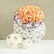 On a bright white tabletop, a beautifully composed centerpiece glows softly against a warm, textured beige backdrop, reminiscent of a calm interior in Clerkenwell's quieter streets. At the top of the arrangement sits a dense, spherical dome of roses in gentle peach and apricot tones. Each rose is fully open and closely nestled against its neighbours, creating a smooth, rounded mound of petals that feels both luxurious and soothing, ideal for a wedding or special dinner near Farringdon. Just below this dome, a ring of white lisianthus blooms fans outward, their petals finely edged in rich purple and violet hues. The colour on the petal rims adds a refined contrast to the soft roses, while fine clusters of white baby's breath are threaded throughout, adding a lace-like lightness and keeping the design from feeling heavy. The blooms are gathered into a low, bulbous container made from intersecting strands of white wicker or rattan, woven into an airy, open lattice that reveals hints of stems and shadow inside. This intricate, handcrafted vessel feels at home among Clerkenwell's design studios and artisan workshops. Resting on the surface in front of the main arrangement are two decorative woven spheres in pale natural wood tones, one slightly larger than the other, echoing the organic curves and textures of the vase. The entire composition exudes understated elegance, natural charm, and a sense of thoughtful design, well-suited to modern wedding tablescapes, intimate gatherings, or styled interiors across Clerkenwell, where contemporary living meets quiet romance.