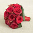 Set against a softly textured beige fabric that resembles natural linen, a compact, rounded bouquet of red roses draws the eye with its luxurious depth of colour and form. The roses are gathered tightly together, each bloom at its prime, layered with plush, velvety petals ranging from vivid scarlet to deep wine red, creating a rich, almost theatrical effect. A few petals curl slightly at the edges, hinting at movement and life, as though the bouquet has just been lifted from a bridal photo session in Clerkenwell and laid gently down for a moment of rest. Nestled between the blossoms, polished red hypericum berries cluster in small groups, their spherical shapes and glossy skins providing a lively contrast against the softness of the roses. Encircling the heads, broad dark green leaves are meticulously folded and smoothed into a firm, decorative collar, their gently reflective surfaces catching the light and giving the whole arrangement a tailored, modern silhouette. The stems are finished with a vivid red satin ribbon that wraps the handle in clean, even layers, suggesting both comfort in the hand and attention to every finishing detail. Photographed from a slightly elevated perspective, the design appears sculptural yet inviting, as if ready to be carried down the aisle of a Clerkenwell townhouse wedding or presented as a passionate anniversary gift in a restaurant near Farringdon. The combination of rich reds, structured foliage, and understated background speaks of classic romance, elegant London style, and the steady, heartfelt craftsmanship of a dedicated local florist.