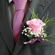 Pinned to a charcoal suit jacket, this blush-pink rose boutonniere offers a tender focal point, like a small promise worn over the heart on a Clerkenwell wedding day. The rose itself is beautifully open yet still composed, its velvety petals curling outward with a satiny sheen, the colour soft enough to feel almost translucent at the edges. Clusters of white gypsophila nestle around the bloom, their tiny, star-shaped flowers creating a misty halo that lightens the composition and contrasts with the darker weave of the lapel. Slender green foliage arcs gracefully outward, adding a sense of movement that recalls the gentle sway of leaves in Spa Fields or along the quieter side streets off Clerkenwell Green. At the base, a narrow ribbon in a muted lilac or mauve tone is wrapped with care and tied into a discreet bow, its texture smooth and slightly reflective, subtly catching the ambient light. The craftsmanship is evident in the careful placement of every stem, the balanced proportions, and the way the boutonniere appears both refined and effortless, as if it naturally belongs with the suit's tailored lines. Seen up close, the details invite touch and imagination: the coolness of the lapel cloth, the softness of the petals, the crisp snap of the stems beneath the ribbon. This is the kind of piece a local florist might prepare for a groom or prom guest heading to a celebration near St John's Square, a small floral artwork that carries with it the joy, nerves, and hope of a day filled with meaningful moments and shared glances.