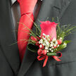 This image shows an impeccably styled boutonniere on a dark charcoal suit, a favourite look for smart occasions around Clerkenwell and nearby Farringdon. At the centre sits a perfectly formed red rose, leaning toward a rich, pinkish-crimson tone that feels warm and inviting even against the sharper lines of the suit. The petals are lush and velvety, opening in a graceful spiral that draws the eye inward, like a quiet focal point amid the bustle of London streets. Surrounding the rose, soft, starry clusters of white baby's breath create a delicate halo, each tiny bloom brightening the composition and lending a sense of airy elegance. Behind and around these, light feathery greenery reaches outward, giving the design a hint of movement, as if stirred by a gentle breeze drifting along Clerkenwell Road. Nestled at the base are small red berries, smooth and glossy, adding a playful glint that echoes the rich hue of the scarlet tie worn above. The stems of the boutonniere are carefully bound with a tidy red ribbon, wrapped smoothly and securely, a subtle sign of meticulous craftsmanship. The overall mood is dignified yet approachable, blending refined masculinity with soft romantic details. One can imagine the faint sweetness of the rose mingling with the green scent of foliage, a subtle counterpoint to the crisp scent of suit fabric and cologne as guests gather for a ceremony or reception nearby. The lighting highlights every curve and texture, showcasing the florist's skill in balancing colour, form, and proportion to create a wearable floral accent that feels like a personal signature of celebration.