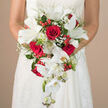 Held just at the waist, this luxurious cascading wedding bouquet seems to flow like a gentle waterfall over the front of a white, finely textured bridal gown, whose lace-like details mirror the intricate beauty of each bloom. Deep crimson roses, fully opened and plush, form the romantic core of the arrangement, their velvety petals curling inward in tight spirals that suggest depth, intensity, and enduring love. Nestled among them are luminous white lilies, their broad, star-shaped petals unfurling with crisp edges and cool satin-like surfaces, accented by fresh green stamens that bring a hint of garden vitality. From the centre, white Phalaenopsis orchids tumble gracefully downward in slender arcs, each bloom silky and luminous, with developing buds along the stems adding natural movement and a sense of unfolding celebration. Wisps of delicate white baby's breath drift between the larger flowers, giving a soft, airy haze that lightens the composition. Glossy red berries, likely Hypericum, add bright sparks of colour and a festive feel, standing out against lush greenery that includes fine, feathery fronds and broader, structured leaves. Small green-tipped white blooms peek through the foliage, offering a quiet nod to detail and expert craftsmanship. The neutral, softly blurred beige background throws the bouquet into sharp relief, so every petal edge, vein, and texture feels almost touchable. The image captures the mood of a sophisticated Clerkenwell wedding, perhaps set around Farringdon or near Clerkenwell Green, and evokes the careful artistry a local florist brings to designing a red and white bridal bouquet that will be carried from ceremony to celebratory reception with grace and poise.