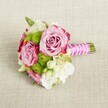 A small, hand-tied bouquet rests horizontally on a softly textured, light beige linen cloth, the neutral fabric echoing the stone tones of Clerkenwell's historic streets. At the centre, three lush pink roses open generously, their petals swirling from a gentle blush at the heart to deeper magenta along the outer edges, creating a natural ombré that feels both romantic and refined. Nestled closely around them are bright lime-green button chrysanthemums, each compact globe made up of tiny, neatly packed petals that add lively texture and a playful contrast to the roses' velvety softness. Threaded between these focal blooms are airy clusters of creamy white flowers, likely lisianthus or freesia, their ruffled petals and slender buds lending lightness and movement, as if the bouquet has just been gathered from a garden near Exmouth Market. Tiny star-shaped white blossoms with soft yellow centres tuck into the gaps, bringing intricate detail and a sense of delicacy. The stems are neatly trimmed and bound into a compact posy, wrapped in a vivid fuchsia satin ribbon that gleams subtly in the gentle, even lighting. Over the ribbon, a fine strand of white faux pearls is crisscrossed in a graceful pattern, giving the handle a touch of bridal elegance that would feel perfectly at home in a wedding at a Clerkenwell town hall or a chic warehouse venue. The overall mood is fresh, modern, and romantic, ideal as a bridesmaid's bouquet, a petite bridal posy, or a charming accent arrangement for engagement celebrations and intimate gatherings across EC1.