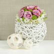 On a smooth, reflective white surface, a meticulously arranged floral centerpiece glows with soft wedding romance, as if placed on a reception table in a chic Clerkenwell loft. The heart of the design is a perfectly rounded dome of flowers, composed of pastel pink roses with hints of deeper blush along their petal edges, and lively lime-green button chrysanthemums nestled between them. The roses are full and velvety, each bloom opening generously, while the small, rounded chrysanthemums provide a fresh, almost playful texture that keeps the composition feeling modern and light. These blooms are gathered into a distinctive spherical vase made from white-painted, intertwined branches, forming an airy, openwork structure that looks both rustic and contemporary-much like the converted warehouse spaces off St John Street. At the base of the arrangement, a gentle skirt of creamy-white blossoms and tight buds drapes softly over the rim, introducing a feathery transition from the lush dome into the sculptural container. To the left, two pale wooden decorative balls, woven into open lattice spheres, continue the theme of natural fibers and gentle curves, staged on the gleaming table as if part of a carefully curated wedding tablescape. The background is a warm, softly textured neutral, blurred enough that it recalls woven fabric or calm plastered walls of a Clerkenwell townhouse, allowing the blush, cream, and green tones to stand out. The whole scene feels serene and polished, well suited for a wedding breakfast near Farringdon, an intimate ceremony by the registry office, or a stylish celebration where flowers add a quiet note of joy.