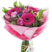 There is an unmistakable glow of joy in this hand-tied pink bouquet, photographed against a bright white backdrop so every petal and texture reads clearly. At its heart, large gerbera daisies in a vivid fuchsia pink command attention, their densely packed, velvety petals curving outward from dark, almost black centers that give the colour a dramatic depth. Interspersed among them are multiple soft pink roses at varied stages - some unfurling in layered spirals, others still tight, glossy buds - which soften the composition with a tender, creamy blush. Small clusters of deep purple-blue statice or similar fillers thread through the stems, offering tiny, papery textures and a cool counterpoint to the hot pinks. Lush greenery - broad, smooth leaves and slimmer stems - frames the blooms and adds a fresh, living backdrop. The bouquet is set into a clear cylindrical glass vase so the bundled stems show through the water line, and a piece of rustic twine is tied around the vase neck holding a small white rectangular tag printed with a stylised pink-and-green rose emblem that hints at a local florist signature. The flowers are wrapped in layered, crinkled sheer pink tissue or cellophane that graduates from a near-translucent pastel halo at the top down to a richer, more saturated pink at the base, creating volume and movement. In soft morning light the arrangement reads as celebratory and intimate - an ideal choice for sending thanks, marking a birthday, or brightening a flat near Exmouth Market or Clerkenwell Green - its scent gentle, sweet, and lingering like a walk past a neighbourhood flower stall.