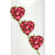 Three sumptuous, heart-shaped floral arrangements created entirely from red and pink roses descend in a graceful cascade across a bright white backdrop, forming a powerful statement of love and remembrance. Each heart is densely packed with roses in varying stages of bloom, from tight, hopeful buds to open, full-petalled flowers, giving the surface a rich, almost velvety texture. The dominant deep crimson tones speak of passion and devotion, while the interspersed fuchsia and blush pink roses soften the palette, adding layers of sweetness and tenderness that feel especially poignant for a farewell. Fine strands of green ivy trace the outline of each heart, their small, fresh leaves offering a natural, gently rambling contrast to the structured shapes. These trailing vines extend between the hearts, visually linking them so they appear as one continuous chain, suggesting the unbroken bond between generations of a family or the enduring connection between partners. The white background, evenly lit and free of distraction, allows every hue and contour to stand out clearly, similar to how such a tribute might appear when placed against a simple wall in a Clerkenwell chapel or memorial space. The lighting is soft but bright enough to highlight the natural sheen of the petals and the crisp edges of the hearts, underscoring the skill and care involved in their creation. As a funeral tribute, this arrangement feels both romantic and respectful, ideal for honouring the life of someone deeply loved and ensuring that their memory is framed in beauty and heartfelt symbolism.