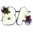 A solemn, hand-crafted pair of freestanding floral initials reading BA, presented against a clean white background. Each capital letter is densely faced with hundreds of small, snow-white chrysanthemum florets, layered so closely they form a pillowy, cloud-like surface that catches soft light and suggests quiet calm. The contours of both the B and the A are crisply defined with a ruffled band of royal blue satin ribbon, the deep blue edge giving the white a strong, respectful frame. At the upper left curve of the B sits a concentrated accent: several velvety deep red roses in mixed stages from tight buds to mid-open blooms, interspersed with narrow, electric-blue delphinium-like spikes that add a cool, vertical contrast. Similar accents appear at the lower right of the A, mirroring the composition and balancing the tribute. Dark green leaves and pale green trailing tendrils weave through the clusters, softening edges and adding an organic fall that breaks the letters' geometry. The two letters are mounted side-by-side on a subtle dark green rectangular base that keeps the display unified and stable for viewing during a funeral service or memorial. Up close you can sense the difference in textures-the roses' velvet and the chrysanthemums' cushiony faces-while the blue ribbon provides a satin sheen under gentle light. This bespoke sympathy piece carries a composed, respectful mood; it reads as a personalised farewell that speaks of remembrance and affection, created with the measured craft of an experienced florist for a memorial setting where colour, form, and sentiment must be held in careful balance.