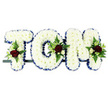 Personalised funeral flower tribute spelling out TOM in three sturdy, block-style capital letters, created as a dignified memorial piece suited to services in Clerkenwell. Each letter is completely covered with tightly clustered white chrysanthemums, the blooms layered close together to form a smooth, plush surface that reads clearly from a distance while still looking soft and comforting up close. Around the outside edge of every letter runs a narrow ribbon of royal blue satin, its clean, lustrous line providing definition and echoing the formal tones often chosen for sympathy flowers. Carefully placed on the 'T', 'O', and 'M' are small, elegant groupings of accent flowers: velvety, deep red roses in full bloom, delicate white or cream spray roses, and graceful white freesia-like stems, all nestled into pockets of rich green foliage and airy ferns. These touches of red and green break the white expanse in just the right places, adding depth, warmth, and a hint of natural movement. The letters rest on a slim, dark support bar that runs unobtrusively behind them, implying that the tribute is ready to be displayed beside the coffin, at the front of a chapel, or in a memorial space. The overall composition feels serene and respectful, like something you might see at a service near Clerkenwell Green or in one of the area's historic churches. It expresses love, remembrance, and gratitude for Tom's life, combining traditional floristry techniques with thoughtful personalisation to help family and friends say goodbye with grace.