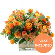 A vibrant arrangement of orange, yellow, and peach-toned flowers is photographed against a bright white background, the colours glowing as if caught in soft midday light over Clerkenwell. At its core are several lush orange roses, their petals spiralling outward with hints of yellow near the centres, creating the impression of small, contained sunsets. Alongside them sit sunny yellow garden roses, more tightly furled yet still full and ruffled, offering a gentle contrast in shape and shade. Woven throughout the bouquet are delicate sprays of peach and light orange alstroemeria, their elongated petals decorated with subtle stripes and speckles, adding an element of fine detail that feels almost like brushwork in a painting. Rising from among these softer forms are numerous small, globe-shaped, spiky orange flowers, reminiscent of safflower or thistle, whose bold texture gives the design a hint of modern, sculptural energy, like public art tucked into a corner of Clerkenwell Green. Surrounding the flowers, an abundance of foliage-smooth, silvery-green eucalyptus leaves and darker green stems-spills outward, creating a lush frame that deepens the overall composition and makes the warm blooms appear even brighter. The entire bouquet is gathered into a clear, square glass vase, where the stems are visible through the water, lending an honest, straightforward feel to the arrangement. Around the middle of the vase, a band of natural beige burlap or jute is wrapped neatly, its coarse weave bringing a rustic, tactile charm to the sleek transparency of the glass. In the lower right corner, a bold orange circle contains the clean white words VASE INCLUDED, indicating that this is presented as a complete, ready-to-place floral gift, ideal for brightening flats, studios, or workspaces across Clerkenwell.