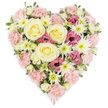 A lush, heart-shaped floral wreath fills the image, set against a pure white background that makes its gentle colours and dense texture stand out. The heart is richly woven from flowers in soothing shades of blush pink, creamy ivory, pale yellow and touches of deeper magenta, all nestled so closely together that the surface looks like a soft, petal-covered cushion. At its centre, large cream-to-light-yellow roses bloom in graceful spirals, their full, layered petals radiating a sense of warmth, respect and enduring affection. Around them, waves of pastel pink carnations form the bulk of the design, their frilled edges overlapping to carve out the heart shape, giving the tribute a comforting, almost embrace-like appearance. Between these, white spider chrysanthemums stretch their slender, spiky petals from vivid green centres, adding brightness, airiness and fine detail that catches the eye. Scattered throughout are clusters of deep magenta-pink lisianthus, their slightly ruffled, bell-shaped flowers deepening the colour palette and lending an undercurrent of poignant emotion. Small flashes of variegated greenery and fresh foliage peek out at the edges, framing the blooms and preventing the arrangement from feeling too heavy. From the pointed tip at the base of the heart, a delicate pink ribbon just peeps into view, quietly indicating that this is a thoughtfully prepared funeral or sympathy tribute. It is the sort of heartfelt arrangement a family in Clerkenwell might choose for a service near St James's Church or along Clerkenwell Road, a floral expression of compassion, farewell and cherished memories shared.