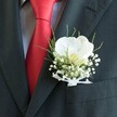 A richly detailed close-up shows the upper torso of a man dressed in impeccably tailored formal wear, as if he is about to step into a wedding venue off Clerkenwell Green. The dark suit jacket, in deep charcoal or black, fits cleanly across his chest, the lapel line crisp and smooth under soft indoor light. Against the jacket, a lustrous red silk tie cascades down in a neat knot, its surface catching subtle highlights that echo the warm glow of an evening reception. Beneath it, a pale, lightly patterned dress shirt brings a gentle contrast, adding texture without distracting from the true focal point: a carefully crafted boutonniere pinned to the left lapel. At the heart of this floral piece sits a single, pristine white Phalaenopsis orchid, its broad petals velvety and luminous, cupping a soft yellow-cream centre that hints at the blush of first light over Exmouth Market. Tiny clouds of white gypsophila, or baby's breath, cluster around the orchid, their miniature blooms creating an airy, almost mist-like halo. Slender strands of fresh, grass-like greenery arc gracefully behind, providing a vivid green backdrop that frames the white flowers. The stems are neatly bound and finished with a delicate sheer white ribbon tied into a small bow, the fabric almost translucent against the dark wool of the suit. The image conveys the quiet anticipation and composed elegance of a groom or prom guest, every detail-a balanced palette of red, white, and green-suggesting thoughtful preparation by a local Clerkenwell florist attuned to both modern style and timeless romance.