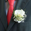 Pinned neatly above a suit pocket, this classic boutonnière embodies the polished, urban romance often found at weddings around Clerkenwell's historic streets. A single white orchid takes centre stage, its petals smooth and almost porcelain-like, catching the light with a gentle sheen. Each curve of the bloom is carefully opened, revealing a soft, creamy surface and a discreet flicker of pale gold at its heart, suggesting warmth and devotion without any need for excess. Surrounding the orchid, a delicate cloud of gypsophila, or baby's breath, spills outward in tiny white clusters, creating a soft, misty halo that feels light as air. Fresh green foliage threads between the blossoms, slender and bright, lending a touch of natural movement that contrasts beautifully with the crisp edge of the pocket. The stems are bound with a small white ribbon, satin-smooth and precisely folded, reflecting the meticulous hand of an experienced florist used to dressing grooms for ceremonies in nearby registry offices and warehouses turned wedding venues. Behind the flowers, the charcoal lapel of the suit offers a deep, velvety backdrop, while a bold red tie cuts through the composition like a line of pure celebration. The interplay of dark fabric, bright tie, and luminous white bloom creates a sense of balance and style, hinting at photographs taken on cobbled lanes and under old brick arches. This boutonnière feels intentionally quiet yet full of promise, designed to be worn close to the heart on a day defined by connection, commitment, and shared joy in the heart of Clerkenwell.
