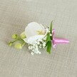 A delicate white orchid boutonniere lies horizontally on a softly textured, oatmeal-coloured linen cloth, its composition captured in gentle, even light that feels like a quiet Clerkenwell morning. At the centre, a single Phalaenopsis orchid bloom opens in pristine white, its broad, velvety petals curving outwards to frame a finely detailed throat touched with soft yellow and the faintest blush of pale pink. Around its base, tiny sprigs of white gypsophila, or baby's breath, cluster like small clouds, their many rounded florets creating a feathery halo that softens the line between flower and fabric. A few slender, fresh green leaves peek out from behind the petals, adding a crisp contrast that hints at the living stem beneath. Extending to one side, a short side stem holds a pair of tightly closed, lime-green orchid buds, their smooth surfaces and pointed tips suggesting blooms still to come, like an unspoken promise on the day of a wedding or prom. The stems are neatly gathered and wrapped in a bright fuchsia pink satin ribbon, its lustrous surface tied into a small, graceful bow with short tails resting lightly against the linen. The ribbon's vivid colour injects a joyful, celebratory note, perfectly suited to a groom's lapel or a special guest's corsage at a ceremony in one of Clerkenwell's historic venues near Exmouth Market or St John Street. The overall impression is one of refined simplicity and quiet elegance, as though the florist has distilled the essence of a modern London celebration into a single, wearable flower.