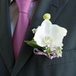 A close-up photograph shows a sharply tailored charcoal or deep black suit jacket, its left lapel in crisp focus, carrying a single white Phalaenopsis orchid boutonniere that immediately draws the eye. The orchid's broad petals are pristine and velvety, curving outwards with sculptural grace, their cool white tone glowing softly against the dark wool. At the flower's centre, a hint of creamy ivory and a touch of yellow-orange in the throat add gentle warmth, like a quiet light in the middle of the composition. Just behind and around the orchid, tiny clouds of white gypsophila and delicate lilac-purple blossoms cluster together, giving the arrangement a soft, misty halo of texture. A small, pale green orchid bud rises just above the main bloom, still closed and full of promise, suggesting new beginnings on a wedding morning in Clerkenwell. The boutonniere is neatly pinned above what appears to be the breast pocket, just beside a sleek tie in rich magenta or deep plum, perfectly knotted over a light grey shirt with a fine, textured pattern. A narrow lavender satin ribbon is wrapped and tied at the base of the stems, its sheen catching the light and echoing the lilac filler flowers, hinting at careful colour coordination often seen at registry office ceremonies off Farringdon Road or intimate celebrations near Exmouth Market. The overall feeling is one of understated luxury and quiet romance: a groom's lapel detail, designed with a florist's practiced hand, evoking soft fragrance, fresh petals, and the poised stillness just before vows are spoken in the heart of London.