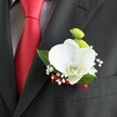 A close-up photograph focuses on a single, meticulously crafted wedding boutonniere, pinned neatly to the lapel of a dark charcoal grey suit jacket with a subtle woven texture, the kind you might see on a groom stepping out of a Clerkenwell town hall ceremony. At the heart of the design sits a large white Phalaenopsis orchid, its wide, velvety petals curving softly outward and drawing the eye to a delicate yellow-and-white lip at the centre. Nestled just above and beside the orchid are two small, rounded lime-green chrysanthemum-style button blooms, their tiny petals forming a tight, almost cushion-like surface that adds a bright, playful contrast. Surrounding these focal flowers, airy sprigs of white baby's breath scatter tiny, cloud-like blossoms that soften the overall outline and bring a feeling of lightness. A few broad, deep green leaves frame the arrangement, their surface catching a gentle sheen of light and grounding the boutonniere in fresh, natural foliage. The base of the stems is wrapped in a slender deep red satin ribbon, carefully wound and secured so that it mirrors the bold red of the solid satin tie just to the left. That tie lies crisply against a light grey and white small-checked dress shirt, the pattern giving extra texture without overpowering the florals. Soft, even lighting highlights the graceful arc of each petal and the fine stitching of the jacket's pocket beneath the boutonniere, suggesting a moment just before the ceremony begins. The whole composition evokes quiet confidence and polished elegance, a refined floral accent ready for a wedding or formal gathering in the heart of Clerkenwell's historic streets.