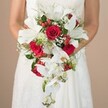 A bride stands tenderly holding a sumptuous, cascading wedding bouquet that radiates romance and celebratory spirit, as seen in countless Clerkenwell nuptials. The bouquet is a lavish blend of large, open white lilies, cascading white phalaenopsis orchids, and rich red roses, beautifully offset by airy clusters of baby's breath, vibrant red hypericum berries, and a mix of wispy green ferns and textured green foliage. Each bloom is placed with florist precision, and the white and crimson tones create a dramatic impact against the softly blurred beige background reminiscent of Clerkenwell's artisan studios. The bride's bare, gently tanned arms and the delicate lace of her gown add a layer of intimacy. This sophisticated arrangement captures both the tradition and modernity found in local celebrations, making it a perfect choice for weddings throughout the area-from creative spaces on St. John Street to cherished historic venues-an emblem of joy, passion, and timeless elegance.