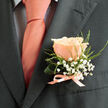This portrait of a boutonnière places a single peach-pink rose at the heart of a quietly elegant story: its petals form concentric layers of soft blush, each one catching a wash of soft daylight so the bloom appears almost luminous against the charcoal-grey lapel. Surrounding the rose, tiny white gypsophila float like a miniature cloud, their delicate clusters and airy stems introducing a lace-like texture that gently softens the suit's tailored silhouette. Wisps of fine green foliage thread through the arrangement, adding movement and a whisper of countryside freshness as though the stem had been clipped at dawn. The base is restrained and refined-a neat knot of satin ribbon in a matching pale peach, its sheen reflecting the warm tones of the wearer's silk tie that shows a subtle dark grid over a pale shirt, tying the palette together with quiet precision. The light in this image is forgiving and natural, revealing the velvet surface of the petals and the tactile contrast between silk, cotton and wool. There is a soft scent suggested here: the barely-there sweetness of a freshly opened rose, intimate and reminiscent of sun-warmed gardens. This small floral accent reads as a gesture-an emblem of celebration, of vows, of formal evenings where detail matters. It's the kind of boutonnière you might see arriving in a brown-paper bouquet from a local Clerkenwell florist for a wedding on a warm spring afternoon, crafted to be felt as much as it is worn.