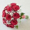 An opulent cluster of richly red roses, ringed by wisps of airy white baby's breath, forms the focal point of this classic bridal bouquet, lovingly arranged by Clerkenwell Flowers for celebrations in EC1 and beyond. The compact, rounded shape showcases each rose's plush, velvety texture, some petals gently unfurling while others remain delicately closed in promise. Between the blooms, tiny white gypsophila creates a floaty, cloud-like contrast and introduces softness to the vibrant display. Striking hints of glossy green foliage are threaded throughout, grounding the composition in nature. At the base, the stems are tightly bound by a wide red satin ribbon crosshatched with elegant white stripes, finished with a coiling strand of small pearls-a refined detail perfect for holding on the walk down a Clerkenwell aisle. Illuminated by bright, natural light that brings forth every nuance of color and texture, this bouquet encapsulates the enduring symbolism of love, commitment, and everlasting joy. It is an ideal choice for weddings at historic local venues or modern celebrations alike, representing the union of tradition and artistry found at the heart of Clerkenwell's vibrant floral scene.