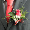 In this close-up boutonnière, a beautifully formed hot pink rose becomes the quiet hero of the occasion, poised against a dark suit that could be worn to a registry office wedding near Farringdon Station or an intimate celebration off Rosebery Avenue. The rose's petals are lush and velvety, folding inwards to a tightly packed heart, then unfurling in gentle spirals that suggest tenderness and devotion. Nestled around the rose are clusters of baby's breath-tiny white florets on slender stems, so light and cloudlike they seem to float, providing a soft contrast in both colour and texture. Wisps of fine, feathery grass arch around the design, creating a sense of movement without overwhelming the compact shape, much like the quiet flow of people through Clerkenwell's cobbled streets on a weekend morning. A single, glossy leaf anchors the back of the arrangement, its deep green surface catching a slight shine that adds structure and balance. At the base, a discreet red ribbon is tied with precision, adding a note of celebration and echoing the rose's warm tones without stealing the spotlight. The whole piece feels thoughtfully proportioned, fresh, and full of gentle romance, as though prepared moments ago at a small studio just off Clerkenwell Road. Ideal for a groom or honoured guest who values subtle beauty and craftsmanship, this boutonnière suggests a day of heartfelt vows, shared laughter, and memories that linger long after the petals have faded.