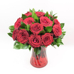 A luxurious bouquet of deep crimson red roses fills the frame, each bloom tightly clustered into a generous, rounded dome that feels both lavish and precise. The rose heads are perfectly formed, with velvety petals spiralling from densely furled centres to more open outer curls, creating a rich tapestry of texture and shadow. Their colour is an intense ruby red, saturated and glowing, the sort of classic romantic hue that immediately suggests whispered promises and heartfelt declarations. Nestled between the roses is an abundance of dark green foliage, likely Ruscus or a similar glossy leaf, its smooth surfaces and pointed tips offering a cool, fresh contrast to the softness of the petals. All of this is gathered into a translucent ruby-red glass vase with a subtle twisted or spiralled pattern running through the glass, catching the light like ripples on the surface of the Regent's Canal on a bright Clerkenwell afternoon. The vase flares gently at the top and rests on a sturdy base, giving the arrangement a confident, grounded presence, while the green stems are faintly visible through the coloured glass, adding depth and structure. The image is set against a clean, white background, making the red and green tones appear even more vivid, as if poised on a pristine counter in a local Clerkenwell townhouse. Bright, even lighting emphasises the immaculate condition of each flower and leaf, suggesting freshly arranged blooms ready for same-day delivery across Clerkenwell and nearby streets. The overall mood is unmistakably romantic and celebratory, ideal for anniversaries, Valentine's Day, intimate dinners, or any moment when someone wants to send a passionate, timeless message of love, appreciation, or devotion.
