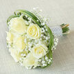 This hand-tied bouquet rests on a neutral, light beige fabric, its diagonal placement creating a graceful, cinematic sweep. Seven prominent creamy white roses anchor the design, their plush petals unfurling in gentle whorls that read as both fresh and timeless. Nestled between those larger blooms are airy clusters of pure white gypsophila, whose tiny stars of flower seem to float and scatter light across the arrangement, lending a cloudlike softness. Woven through and around the flowers are long, glossy loops of green foliage-bear grass by appearance-that sculpt the silhouette and add verdant contrast to the pale palette. The stems are bound tightly and clad in a lustrous, silvery-white satin ribbon; sewn into that wrap are multiple rows of delicate pearl beads that catch the light like tiny memories. Soft, even lighting enhances the cream and ivory tones while preserving the tactile detail of linen beneath, and the whole arrangement suggests the faint, clean scent of roses. This bouquet reads as an emblem of refined celebration-perfect for a bride seeking classic elegance at a Clerkenwell chapel, an intimate registry office, or a spring ceremony in nearby Spa Fields. It could also be offered in quieter moments of sympathy, where its quiet beauty communicates comfort. The composition shows considered florist craft: balance of scale, texture, and movement, with a mood that is at once serene, celebratory, and quietly luxurious.
