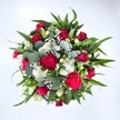 Viewed from directly above against a clean white background, this abundant bouquet forms a near-perfect circle of colour and texture, as if it has just been set down on a white table in a Clerkenwell studio. Deep crimson red roses sit at the heart of the arrangement, some still held in tight, velvety buds and others beginning to unfurl their layered petals, suggesting tender romance and quiet elegance. Around them, rich fuchsia and ruby red carnations ruffle and fold into one another, their plush, frilled blooms adding a joyful, almost celebratory energy. Threaded between the warmer tones are clusters of small, daisy-like white chrysanthemums, their bright centres and crisp petals bringing clarity and light to the design. Scattered delicately through every gap, tiny white gypsophila flowers-baby's breath-create a soft, misty veil, as though the bouquet has been dusted with fine, floral snow. The flowers are cradled by an artful mix of greenery: strong, pointed dark green leaves, likely from lilies or alstroemeria stems, give the arrangement height and structure, while round, silvery eucalyptus leaves add a contemporary softness and a hint of cool blue-green. The lighting is even and natural, with no harsh shadows, allowing every tone and texture to be clearly seen, from the velvety rose petals to the smooth, waxy leaf surfaces. The bouquet feels generous and hand-crafted, the kind of arrangement chosen to celebrate an anniversary on a quiet street off Exmouth Market or to send a heartfelt get well soon to a friend working in one of Clerkenwell's design studios. It radiates affection, vitality, and the simple joy of receiving fresh flowers delivered straight to the door.