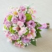 Resting on a pale, textured fabric not far from the creative bustle of Clerkenwell Road, this expertly arranged bridal bouquet radiates joyful elegance. Mauve-lavender roses form the bouquet's plush focal point, their soft curves and intricate petal structure conjuring a sense of romance and promise. Juxtaposed with these classic blooms are distinctive, spherical lime-green button chrysanthemums, injecting playfulness and contemporary flair that echoes the district's lively spirit. Pink Alstroemeria lilies nestle gracefully, their petals delicately adorned with streaks of magenta and flecks of soft yellow, lending optimism and artistic energy to the design. White gypsophila, scattered like airy clouds throughout, infuses the bouquet with softness and ethereal lightness, while lush green leaves peek from beneath, evidencing the freshness and expertise of Clerkenwell Flowers. The entire bouquet is unified by a luxurious fuchsia satin ribbon, meticulously wrapped and enhanced with a delicate necklace of glossy white pearls-an elegant detail fit for special moments. Designed to be cradled by a bride or presented as a gift of gratitude, this arrangement captures the magic of Clerkenwell's community, artistry, and love of celebration, making every occasion memorable and heartfelt.