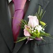 An air of refined celebration lingers in this vivid Clerkenwell image: a close look at a gentleman's charcoal-grey suit set against the urban pulse of Farringdon, featuring a masterfully arranged boutonniere at its heart. The soft blush-pink rose, so delicately unfolded, sits paired with a striking pink-and-purple striped alstroemeria, drawing the viewer's gaze. Tiny clouds of white gypsophila and a bold green chrysanthemum nestle in between, their textures and forms reminiscent of spring blossoms found along Clerkenwell Road. An exquisite bow fashioned from light lavender satin ribbon tucks all elements snugly together, echoing the lively magenta of the silk tie. Lacy ferns and long, fine blades of foliage encircle the blooms, giving every detail movement and depth. The careful craftsmanship and subtle colour harmony reflect both contemporary taste and timeless tradition, making this boutonniere a statement piece for any special occasion in the heart of Clerkenwell.