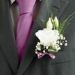 A close, intimate view of a groom's charcoal grey suit in Clerkenwell shows the quiet drama of a single white lisianthus buttonhole against fine formal tailoring. The jacket, likely a smooth wool with a subtle weave, fills most of the frame, its lapel perfectly pressed and catching a soft, even light that feels like a calm wedding morning off St John Street. At the centre of the boutonnière, one fully open lisianthus bloom glows creamy white, its ruffled petals layered like delicate silk, giving a sense of purity and new beginnings. Just above it, two slender ivory buds lean gently forward, not yet unfurled, suggesting the promise of the day ahead. Around this trio, tiny clouds of white gypsophila drift outward, their pinpoint blossoms adding texture and a hint of romance. Fine blades and feathery fronds of greenery arc in light, airy lines, framing the flowers without overwhelming them, and creating a sense of movement against the matte charcoal cloth. Anchoring the design, a narrow mauve ribbon is wrapped neatly around the stems, its sheen echoing the deep magenta or fuchsia silk tie that cuts a bold line between the lapels. The tie is immaculately knotted, its smooth surface contrasting with the patterned white and grey shirt underneath. Together, flower and fabric tell a story of thoughtful preparation and refined taste, as if the wearer were about to step from a townhouse near Exmouth Market into a sunlit ceremony, carrying with him a whisper of fresh, green fragrance and the gentle tension of a day filled with hope.