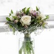 A serene white rose bouquet stands in a clear cylindrical glass vase, set against a luminous, softly blurred white backdrop that suggests a bright Clerkenwell morning filtering through a window. Three creamy white roses, fully open yet still perfectly fresh, form the heart of the design, their velvety petals gently unfurling to reveal subtle ivory centers that speak of purity and new beginnings. Around them, rich emerald foliage curves and folds, with broader leaves cupping the blooms as if protecting them, giving the arrangement a well-balanced, rounded silhouette. Tiny clouds of white gypsophila, or baby's breath, drift between the roses and greenery, their delicate, star-like flowers catching the light and adding a misty, romantic texture. Slim alstroemeria stems, some in bud and some just beginning to open, introduce gentle touches of muted purple and blush-pink, their narrow petals and streaked interiors creating a quiet play of colour that never overwhelms the calm white palette. Through the vase, the neat green stems can be seen, trimmed precisely and gathered with a discreet ribbon above the line of crystal-clear water, a detail that reflects careful, professional craftsmanship. The light streaming from behind highlights every curve and shadow, turning the bouquet into a softly glowing focal point, as if it were placed on a windowsill of a Clerkenwell townhouse overlooking Exmouth Market. The overall feeling is one of peace and thoughtful elegance, perfectly suited to moments that call for a gentle, comforting gesture-perhaps a message of sympathy, a simple thank you, or a gift to bring calm and grace into a quiet London home.