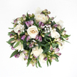Photographed from directly overhead, this hand-tied sympathy bouquet appears like a calm, rounded island of flowers resting on a spotless white surface, every stem and petal clearly visible. At its centre, generous white roses unfurl with quiet confidence, some in full bloom with layered petals revealing creamy ivory and the softest hint of peach, others still more contained, cupped in gentle spirals that feel almost protective. Winding among them, purple alstroemeria buds introduce a cooler tone, their slender, tapering shapes brushed with lavender, violet, and muted green. Most buds remain closed, a few just splitting at the tips to suggest the fine, striped petals within, adding a sense of forthcoming life rather than overt showiness. Sprinkled throughout, clouds of white baby's breath bring an ethereal lightness, each cluster a froth of tiny blossoms that softens the spaces between the larger flowers. Surrounding this is a collar of rich greenery, the deep green leaves smooth, slightly glossy, and gently arched, giving the design structure and a natural frame. The palette of creamy whites, soft purples, and verdant greens feels quiet and meditative, evoking the hush you might find in a tucked-away Clerkenwell side street at dusk. Though the image is still, you can almost imagine the subtle, fresh scent rising from the bouquet. It is the sort of arrangement chosen when you need to send a tender message of support, perhaps delivered from a local Clerkenwell florist to a home near Rosebery Avenue, where it can rest on a side table or mantel, offering a gentle, living symbol of love, memory, and peace.