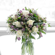 This image shows a tranquil sympathy bouquet arranged in a clear glass cylinder vase, the sort of thoughtful design a local Clerkenwell florist might prepare for delivery to a family in need of gentle support. At its heart are several creamy white roses, fully formed yet soft at the edges, their petals opening in calm, rounded layers that catch the natural light and create a sense of quiet grace. Intertwined with the roses are alstroemeria blooms and buds in shades of purple, mauve, and soft lavender, their slender petals marked with hints of green, bringing a cool, measured touch of colour to the arrangement. Delicate sprays of white baby's breath are scattered throughout, their tiny blossoms forming fine, airy clusters that soften the lines between each flower and lend a light, cloud-like texture. Surrounding everything is lush green foliage, leaves polished and vibrant, some curving gently outward, others standing more upright, providing depth and a feeling of steady support to the paler blooms. The clear vase reveals neatly gathered green stems submerged in fresh water, reinforcing the sense of cleanliness and care. The background is bright and softly white, suggestive of a sunlit interior, perhaps a window looking over the quiet streets and courtyards around Clerkenwell Road, where light filters in gently rather than glaring. The overall effect is calm, respectful, and composed, an arrangement intended to express sympathy, remembrance, and a message of peace, offering visual comfort in times when words may come slowly or not at all.