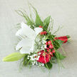 Cradled as though in careful hands in a Clerkenwell studio, this hand-tied wedding bouquet feels like a personal vow made in flowers. A pristine white lily sits proudly at the centre, its broad, creamy petals opening in a calm, star-like shape that radiates purity and hope. The light seems to catch on its satiny surface, revealing the soft texture and the subtle dusting of pollen deep inside the bloom. Encircling the lily, classic red roses give the composition a heartbeat of emotion-their layered, velvety petals unfurling with the richness of true love, long commitment, and quiet passion. Dotted amongst them, clusters of bright red berries, likely hypericum, add cheerful pops of colour, their rounded forms shining like tiny jewels against the deeper shades. Wisps of delicate gypsophila weave through the bouquet, creating airy pockets of white that soften the intense reds and echo the innocence of wedding vows exchanged in nearby churches or registry offices off Clerkenwell Road. Slender sprays of soft greenery frame the flowers, their feathery edges adding lightness and a gentle, natural movement, as if a breeze from the gardens near St John's Square had brushed past. The stems are gathered tightly and bound in smooth scarlet ribbon, the satin wrap neatly finished to give the bouquet a polished, ceremonial feel, perfect for walking down an aisle or posing for photos on cobbled side streets. From its imagined fragrance-a blend of fresh, sweet, and green notes-to its harmonious palette, the bouquet carries a mood that balances joyous celebration with tender reflection, ideal for anniversaries, intimate weddings, or simply saying, You're cherished, with local delivery across Clerkenwell.